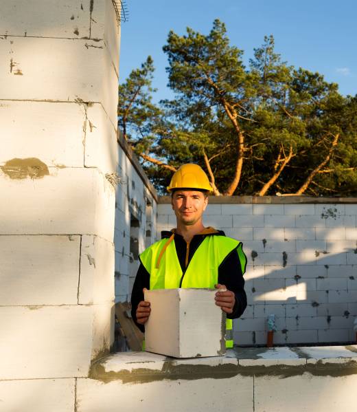 The builder holds a block of cellular concrete in his hands - the masonry of the walls of the house. Construction workers in protective clothing-a hardhat and a reflective vest.
